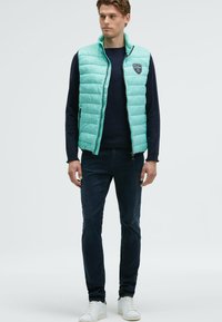 Giovane uomo che indossa un gilet trapuntato color turchese sopra una maglietta nera a maniche lunghe, jeans scuri e sneakers bianche, in piedi davanti a uno sfondo semplice.