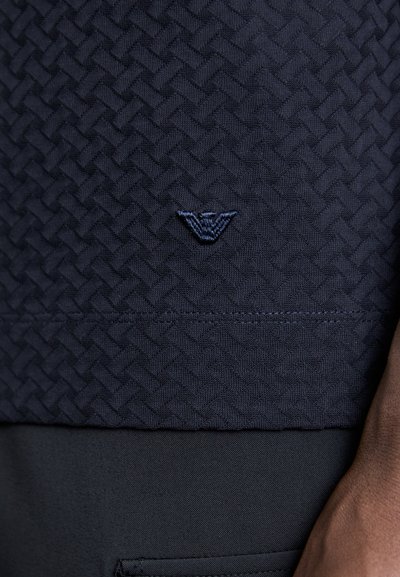 Μπλέ υφασμάτινο πουκάμισο με υφή που διαθέτει ύφασμα ύφανσης, με κεντημένο λογότυπο σε σκούρο μπλε στην άκρη και λείο ύφασμα από κάτω.