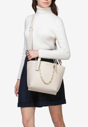 Beige leather handbag with a geometric shape, featuring gold chain accents and dual handles, worn on a shoulder with an adjustable strap.