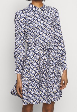 Persona che indossa un vestito a maniche lunghe con un motivo geometrico circolare in blu, bianco e giallo, con cintura in vita e gonna svasata.