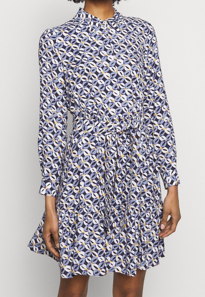 Personne portant une robe à manches longues avec un motif circulaire géométrique en bleu, blanc et jaune, avec une ceinture à la taille et une jupe évasée.
