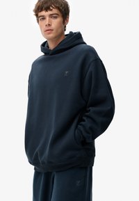 Sudadera azul marino hecha de tejido suave, que cuenta con un bolsillo frontal y un logo en el pecho. Diseñada con un corte relajado y puños acanalados.