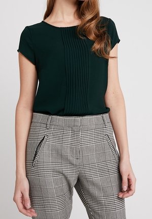 Femme portant un chemisier noir à manches courtes avec des plis verticaux, associé à un pantalon taille haute à carreaux gris et blanc avec poches zippées.