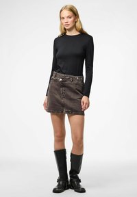 Junge Frau mit langen blonden Haaren, die ein schwarzes Langarmoberteil, einen braunen Jeans-Minirock und schwarze Stiefel bis zu den Knien trägt, steht vor einem weißen Hintergrund.