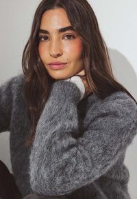 Suéter gris y suave con un diseño texturizado y puños acanalados. La modelo posa con una mano en el cuello frente a un fondo neutro.