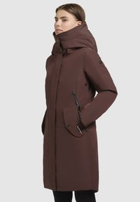 Lang brun parka med høj krave, sidelommer med lynlås og knaplukning foran. Lavet af glat stof med en tætsiddende silhuet og sorte accenter.