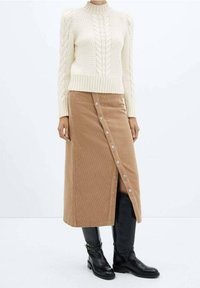 Pull en maille torsadée crème avec ourlet et manches côtelés, associé à une jupe en velours côtelé beige avec une fente latérale et des boutons métalliques, porté avec des bottes noires montant jusqu'aux genoux.