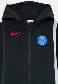 Veste noire zippée avec capuche, swoosh rouge de Nike sur la poitrine gauche et logo bleu du club Paris Saint-Germain sur la poitrine droite.