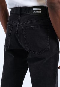 Los jeans de mezclilla negra presentan un corte ajustado, dos bolsillos traseros y una etiqueta de marca en la cinturilla. La textura parece suave con un ligero desvanecimiento.