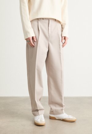 Person trägt beige weit geschnittene Hose, weiße Sneakers mit Gummisohlen und ein cremefarbenes Langarmshirt, steht auf grauem Boden vor einer weißen Wand.
