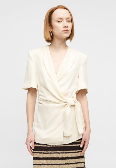 Μπεζ μπλούζα τύπου wrap με κοντά μανίκια και λεπτομέρεια δέσιμου μπροστά. Διαθέτει βαθύ V-λαιμό και ομαλή, υφή υφάσματος.