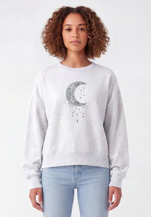 Junge Frau mit lockigem Haar trägt ein hellgraues Sweatshirt mit einem kunstvollen Halbmond-Design und hängenden dekorativen Ketten und Monden.