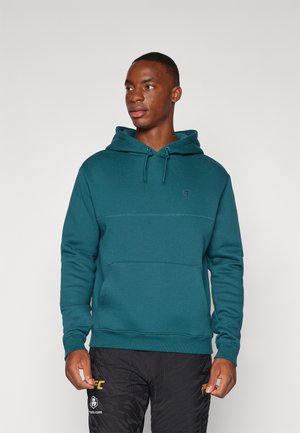 Sudadera verde azulado con un bolsillo tipo canguro, capucha con cordón y un detalle de logotipo sutil. Textura suave y corte relajado. Combinada con pantalones negros con patrón.