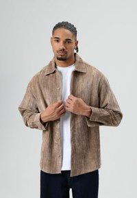 Brown corduroy shirt with a collared neckline and button closure. Soft texture, relaxed fit, and long sleeves. Worn over a white t-shirt.
