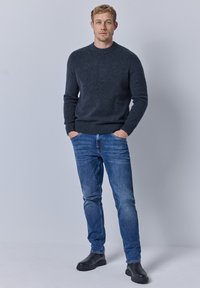 Dunkelgrauer, gestrickter Pullover mit Rundhalsausschnitt, kombiniert mit blauen Jeans. Mann steht mit den Händen in den Taschen und trägt schwarze Lederstiefel.