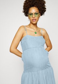 Vestuário sem mangas em chambray azul claro com bainha em camadas, bolso frontal e corte descontraído. Modelo usa óculos de sol verdes e joias em ouro em camadas.