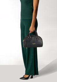 Borsa in pelle nera con parte superiore arrotondata, manici doppi e logo dorato. Il modello indossa un outfit verde e scarpe col tacco nere, tenendo la borsa.