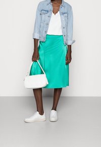 Donna che indossa una giacca di denim chiaro, un top bianco a scallop, una gonna midi turchese, sneakers bianche e tiene in mano una borsa intrecciata bianca.