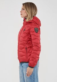 Donna in giacca a cappuccio trapuntata rossa con zip nera e toppe sulla manica, in posizione laterale su sfondo bianco.