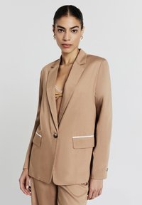 Blazer marron clair avec une texture lisse, fermeture à un bouton et accents blancs sur les poches. Présente une coupe classique et des revers à cran.