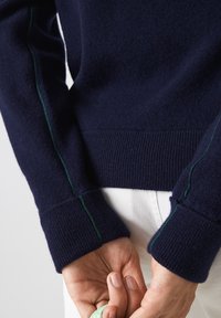 Navy knitted sweater featuring green accent stitching, ribbed cuffs, and a soft texture, partially shown with hands holding a green object.