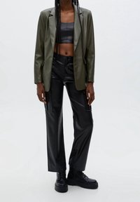 Blazer en similicuir vert olive sur un top cropped noir ; pantalon cargo en similicuir noir ; bottes noires épaisses ; texture lisse, design structuré.