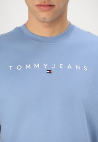 T-shirt di cotone azzurro con ricamo bianco "TOMMYJEANS" e un piccolo logo in rosso, bianco e blu navy al centro.