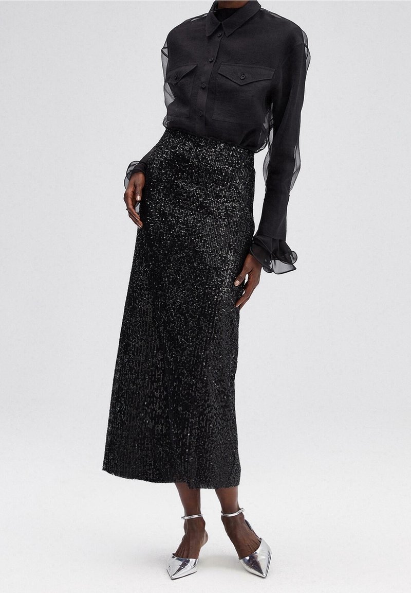 Modèle portant une chemise noire transparente à manches longues avec des poches à boutons et une jupe midi noire à sequins taille haute avec des talons pointus argentés.
