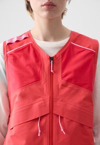 Rote Weste mit durchgehendem Reißverschluss, zwei Brusttaschen und pinken Akzenten. Hergestellt aus leichtem Material, mit einer glatten Textur und modernem Schnitt.