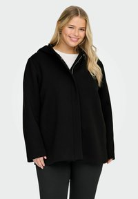 Veste noire à capuche avec une coupe décontractée, fabriquée en matière douce. Elle présente une fermeture éclair à l'avant et des manches longues. Design simple et élégant.