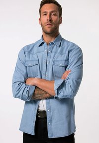 Hellblaue Jeanshemd mit Knöpfen, zwei Brusttaschen, silbernen Knöpfen und hochgekrempelten Ärmeln. Kombiniert mit schwarzen Hosen und einem schwarzen Gürtel.