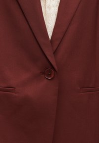 Bordeaux rode blazer van gestructureerde stof met een reverskraag, twee zakken en een enkele knoopsluiting. Witte kanten binnenlaag zichtbaar.