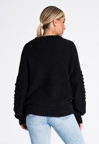 Czarny sweter wełniany o luźnym kroju, z teksturowanymi rękawami i ściągaczem u dołu. W połączeniu z jasnoniebieskimi dżinsami.