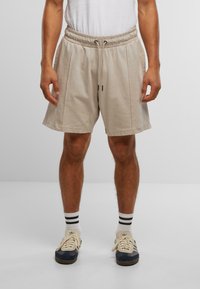 Beige Shorts mit elastischem Bund, Kordelzugdetails und Seitentaschen, aus weichem Stoff. Getragen mit weißen und schwarzen gestreiften Socken.