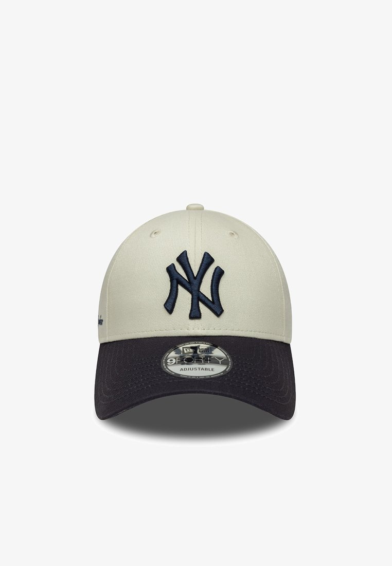Beige baseball cap med en mørk marinblå skygge. Har et forhøjet logo i marinblå på forsiden. Justerbar rem bagpå. Lavet af stof med en glat overflade.