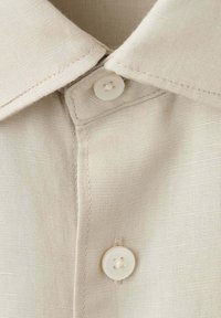 Col de chemise en coton beige avec deux boutons blancs fixés sur la patte de boutonnage, coutures détaillées visibles sur le tissu.