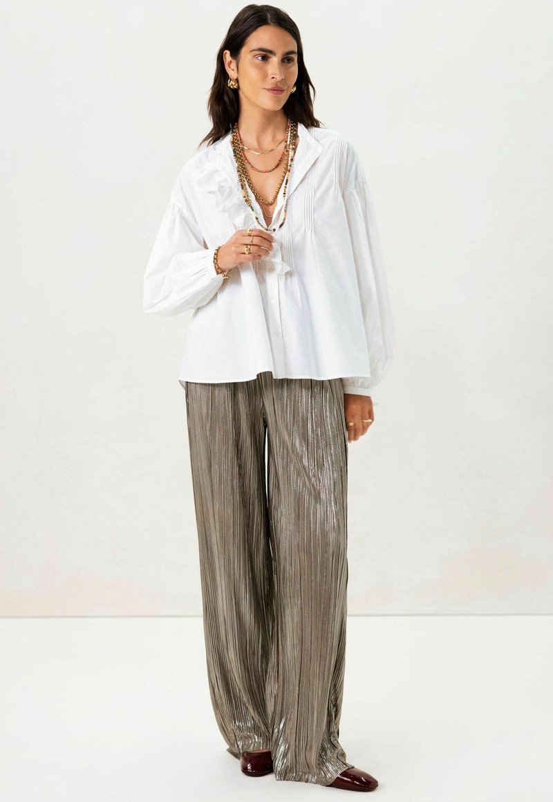 Witte blouse met gerimpelde details, gecombineerd met losse, metallic geplooide zilveren broek. Accessoires omvatten gelaagde kettingen en gouden oorbellen.