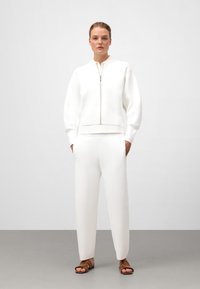 Un sweat à zip blanc et un pantalon assorti en matériau doux, offrant une coupe décontractée, des poignets côtelés et des poches latérales. Des sandales marron complètent le look.
