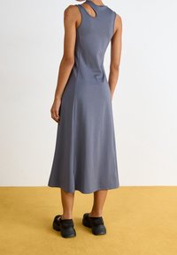 Robe sans manches grise avec un col rond et un détail découpé dans le dos, présentant un design fluide mi-long et un tissu doux. Chaussures noires.