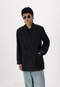Double-breasted black pinstripe coat with wide lapels, textured wool fabric, side pockets, and a relaxed fit over light blue jeans.