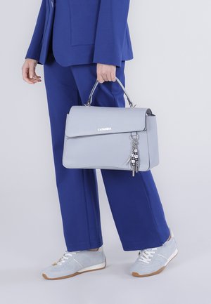 Persona in completo blu che tiene una borsa grigio chiaro con manico in metallo e portachiavi decorativo, indossando sneaker grigio chiaro con lacci bianchi.