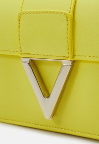 Geel neplederen handtas met een zilveren driehoekige hardwareaccent en zichtbare stiksels. Glad oppervlak, compacte rechthoekige vorm.