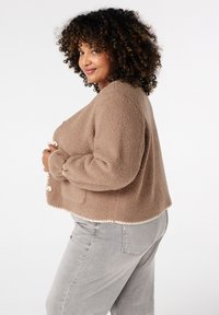 Kruidige vrouw met krullend haar, gekleed in een bruine textuurjacket en lichtgrijze jeans, die zijwaarts glimlacht tegen een effen witte achtergrond.