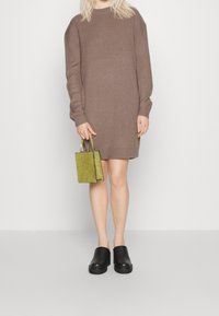 Abito a costine marrone con maniche lunghe, zoccoli neri e una piccola borsa verde texturizzata. Sfondo semplice, focus sull'outfit.