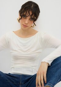 Vit långärmad topp med en rundad halsringning och samlad struktur; matchad med blå denimjeans. Guldfärgade hoops örhängen accentuerar outfiten.