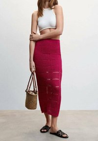 Gehäkelter Midirock in Fuchsia mit geometrischen Aussparungen, kombiniert mit einem weißen, gerippten Crop-Top und schwarzen Sandalen, dazu eine geflochtene Handtasche haltend.