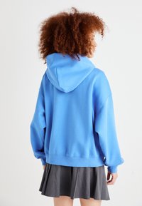 Blå oversized hettegenser i mykt materiale, med kengurulomme, ribbestrikkede mansjetter og kant. Kombinert med et grått plissert skjørt.