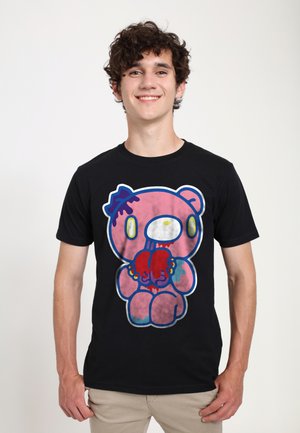 Ung mand med krøllet hår iført en sort t-shirt med en lyserød tegneseriebjørn, der holder et rødt dryppende hjerte.