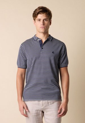 Jeune homme portant un polo à manches courtes rayé bleu marine et blanc et un pantalon beige clair sur un fond beige uni.