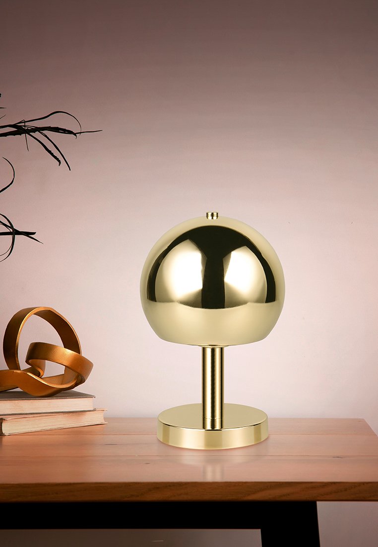 Goldene, metallische Tischlampe mit einem sphärischen Schirm, zylindrischem Sockel und glatter Oberfläche. Auf einem Holztisch platziert, neben dekorativen Gegenständen.